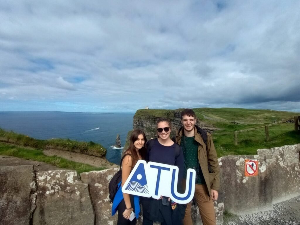 Three International Student Society members holding an ATU sign at the Cliffs of Moher, with the ocean and green cliffs in the background.